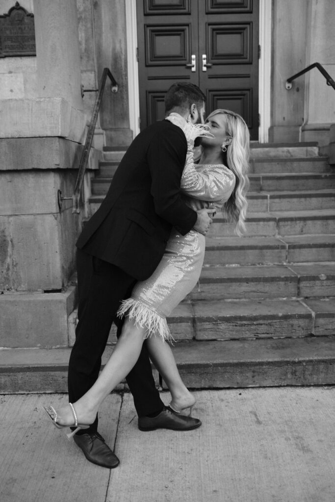 Black and white vertical photo of engaged couple kissing as he dips her in front of stairs at the Niagara on the lake Court house