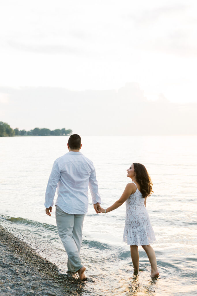 Engaged couple dressed in white at sunset walking along the beach in Niagara on the lake